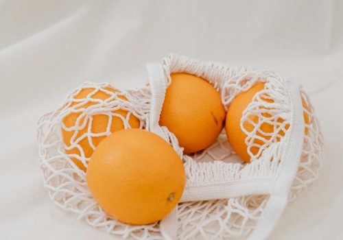Four ripe oranges in an eco-friendly mesh bag on a clean white background.
